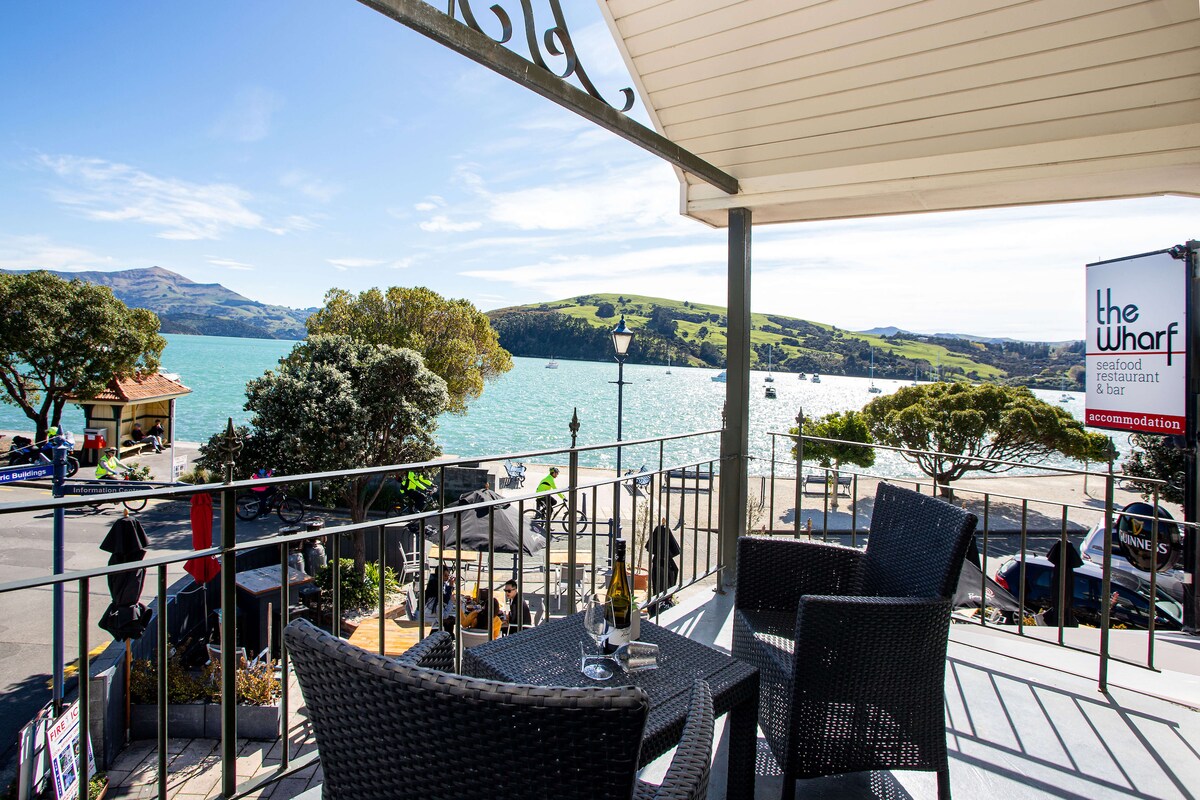 A balcony offers stunning views of Akaroa Harbour, framed by lush hills and a clear blue sky. Outdoor seating is arranged on the balcony, providing a comfortable space to enjoy the picturesque surroundings.