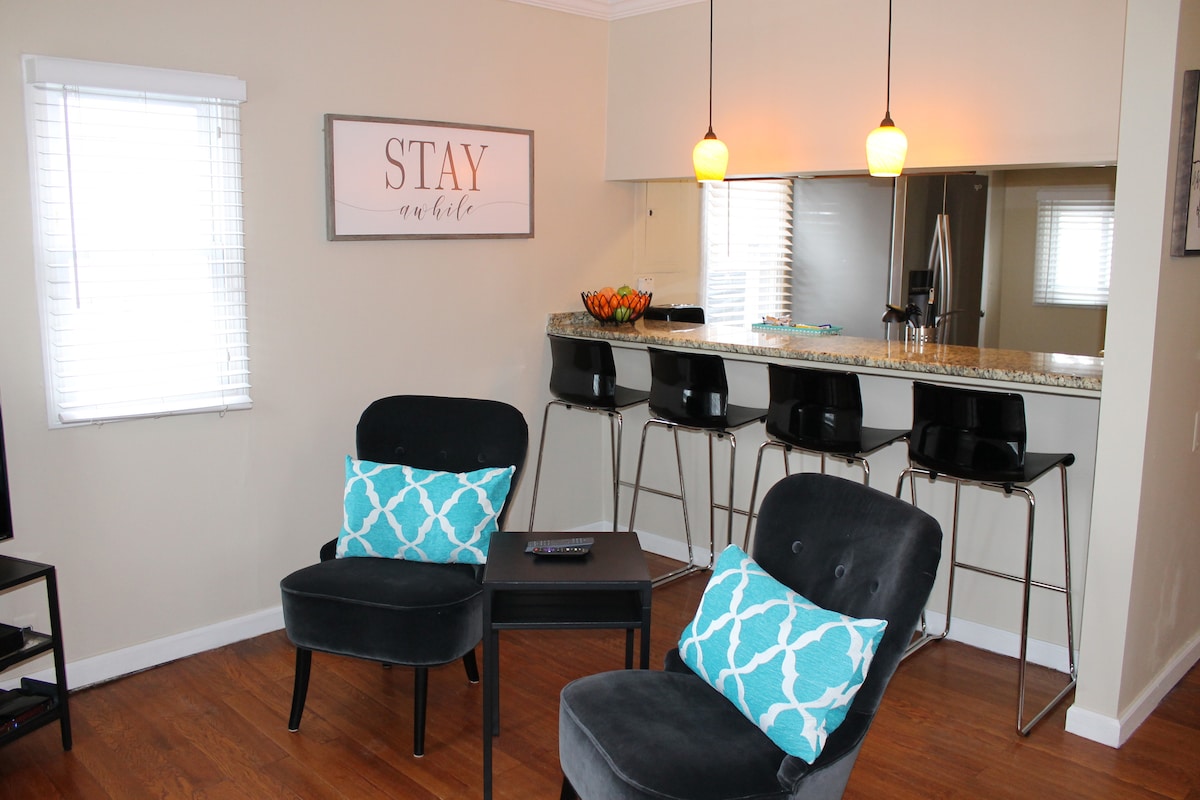 A chic living space is highlighted by two black chairs with turquoise accents, positioned beside a small table. A counter with bar stools is visible in the background under pendant lighting. Natural light streams in through two windows with white blinds.