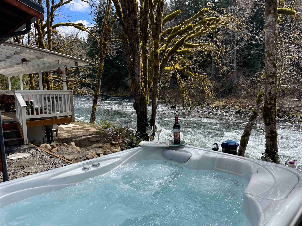 A hot tub is positioned near a river, surrounded by trees with mossy branches. The bubbling water reflects sunlight, while a bottle and glasses rest on the edge. The serene landscape features a view of the flowing river and a nearby pathway leading to the cabin.