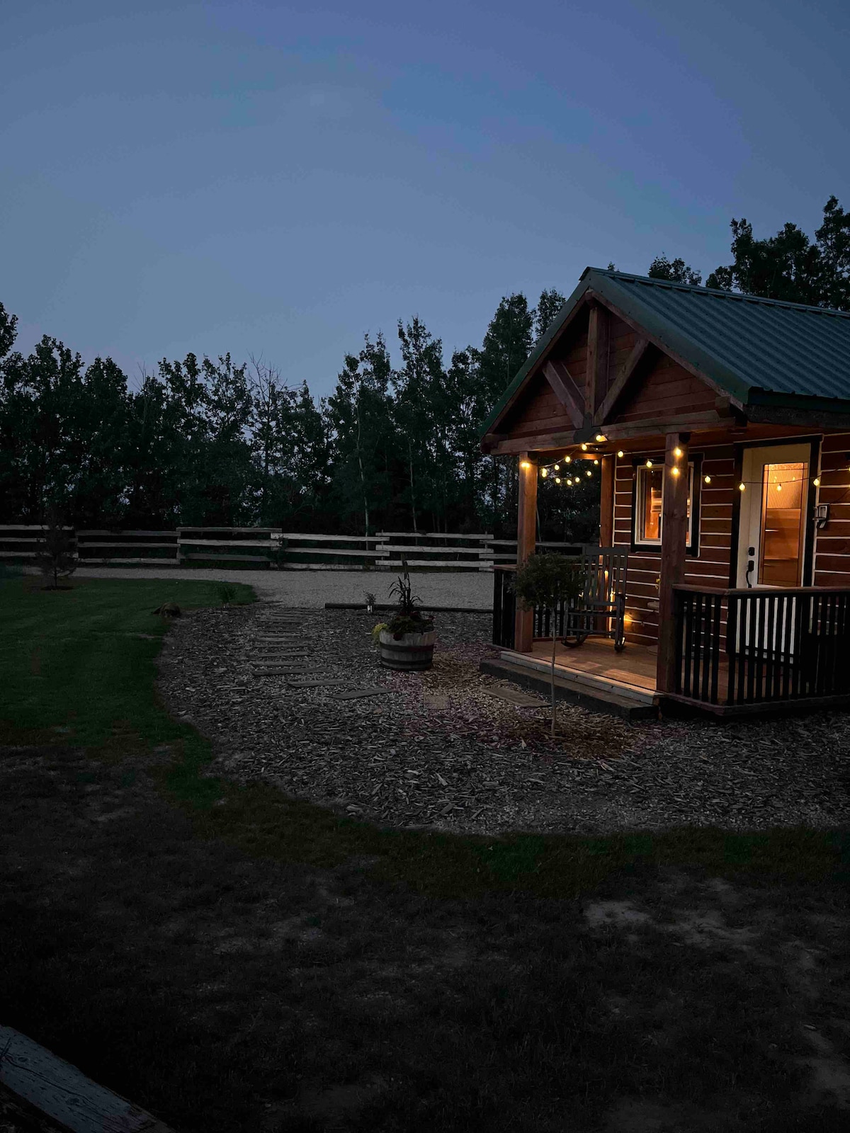 The cabin is softly illuminated by string lights at dusk, highlighting its rustic charm. A gravel area surrounds the cabin, and a wooden fence encloses the property. Trees create a natural backdrop, adding to the serene ambiance of the outdoor environment.