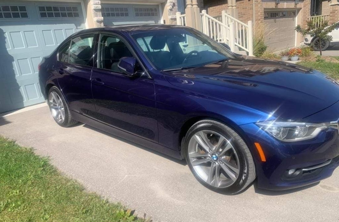 A blue sedan is parked on a driveway, showcasing its sleek design and polished alloy wheels. The vehicle's clean body reflects sunlight, while the garage door and foliage create a welcoming residential scene.