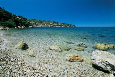 The image presents a clear coastal scene featuring a rocky shoreline. Shallow waters reveal smooth stones and pebbles, transitioning to deeper blue hues as they extend towards the horizon. A tranquil backdrop displays a lush hillside, enhancing the natural beauty of the coastal environment.