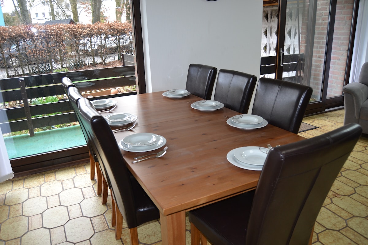 A spacious dining table with eight dark leather chairs is set for a meal. Plates and cutlery are neatly arranged on the surface. Large windows allow natural light to fill the room, while a view of the greenery outside adds to the inviting atmosphere.
