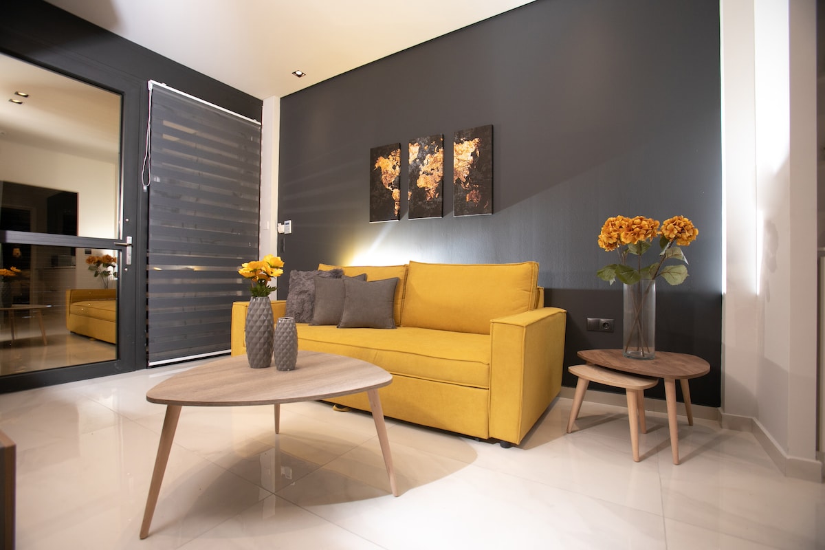 A cozy seating area features a yellow sofa accompanied by gray and textured pillows. Two round wooden tables are positioned in front, with vases of yellow flowers adding a touch of color. Natural light filters in from the adjacent glass door, illuminating the modern decor.