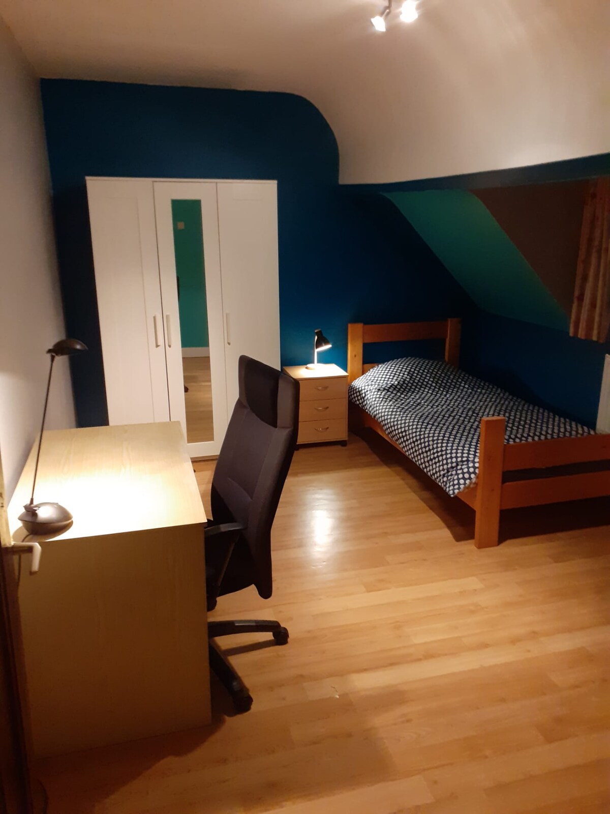 A private room is shown with a single bed positioned against a blue wall. A wooden desk and ergonomic chair are nearby, alongside a wardrobe with mirrored doors. Soft lighting enhances the calm, spacious feel of the room, which features wooden flooring throughout.