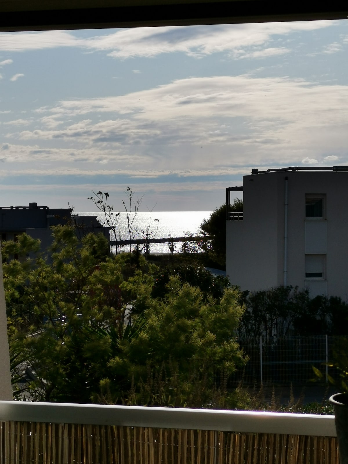A view from the terrace overlooks the ocean, with shimmering water reflecting the sunlight. In the foreground, trees and greenery create a natural frame, while a few buildings are visible in the background, contributing to the coastal ambiance.