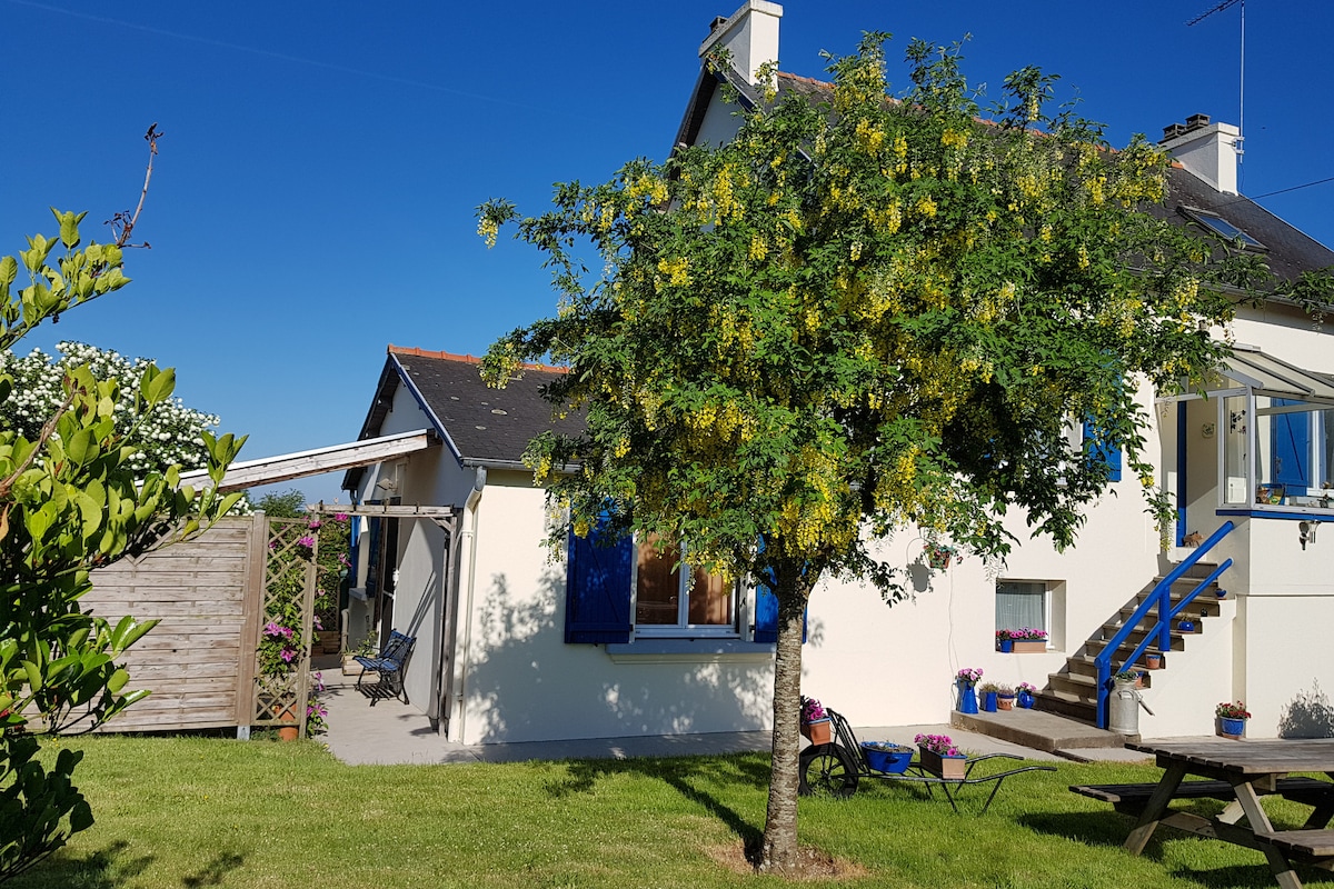 A charming countryside gîte is surrounded by lush greenery and features a large tree in the foreground. The exterior showcases a mix of white and blue elements, with a staircase leading to an upper level. Bright flower pots add color along the base of the building.
