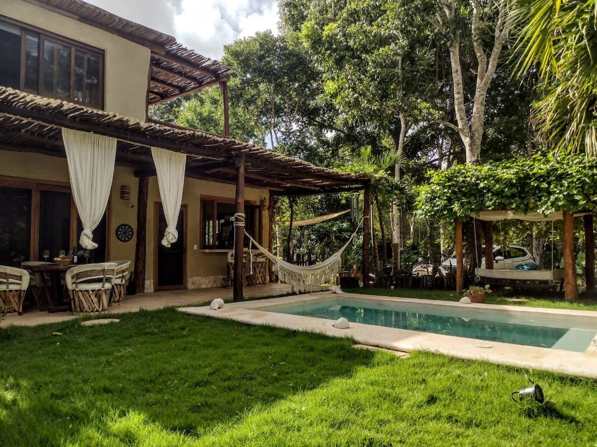 A serene outdoor space features a refreshing pool surrounded by lush green grass. A shaded area with hammocks offers a relaxing spot, complemented by wooden seating. The villa's exterior is framed by trees, providing a peaceful and private atmosphere.