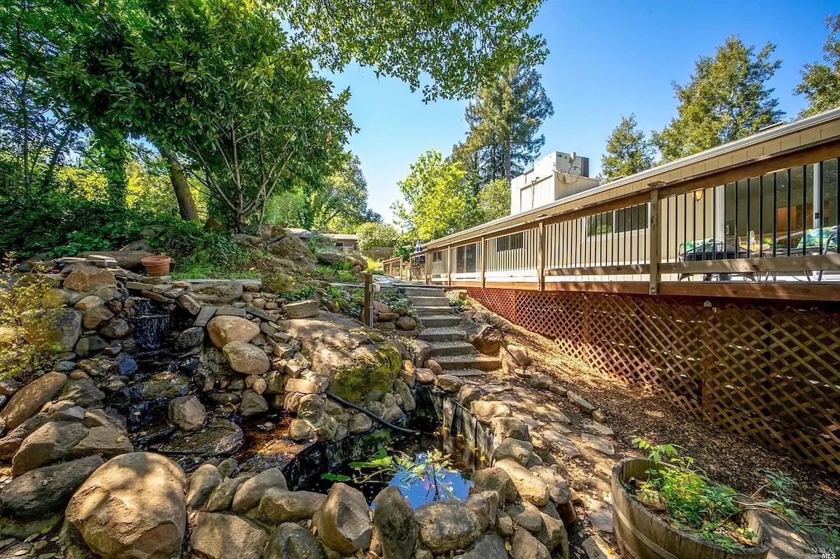 A tranquil outdoor setting features a natural rock pond, bordered by an arrangement of varied stones and lush greenery. A wooden deck extends along the home, providing access to the indoor spaces. Bright sunlight filters through the tree canopy, highlighting the serene atmosphere.