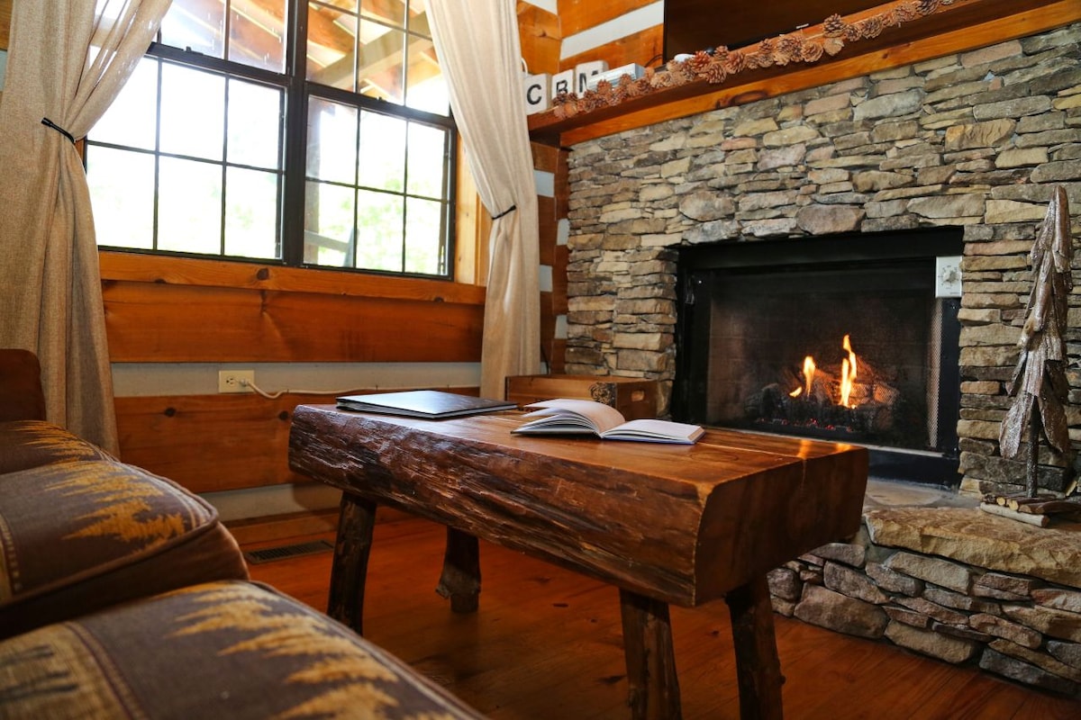 A rustic living space features a stone fireplace with a crackling fire, providing warmth. A wooden coffee table, crafted from natural materials, rests in front of a cozy sofa. Soft curtains frame the window, allowing natural light to brighten the room.