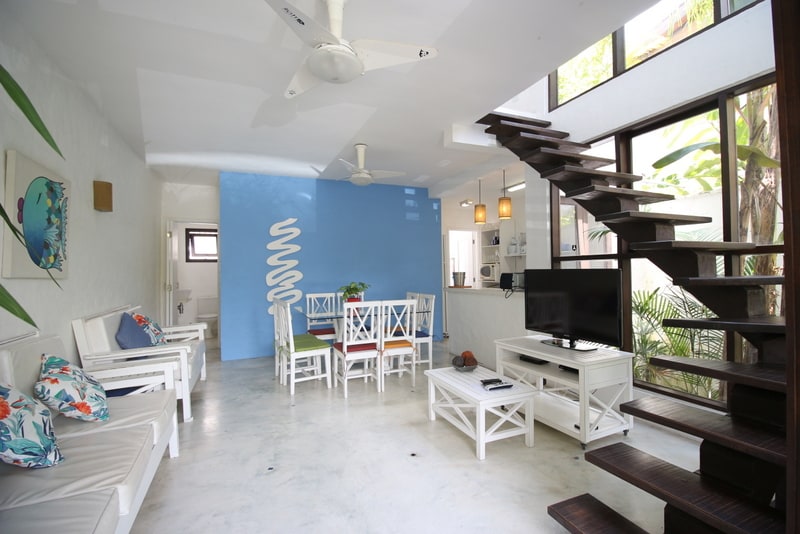 A spacious living area features a light color palette, complemented by a vibrant blue wall. White seating arrangements provide comfort, while a television stands on a white media console. A staircase leads to an upper level, surrounded by lush greenery.