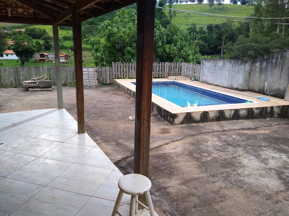 An outdoor area features a rectangular swimming pool surrounded by a tan border. Green hills and trees are visible in the background. A wooden post and a stool are positioned in the foreground, and a small grassy area is also present.