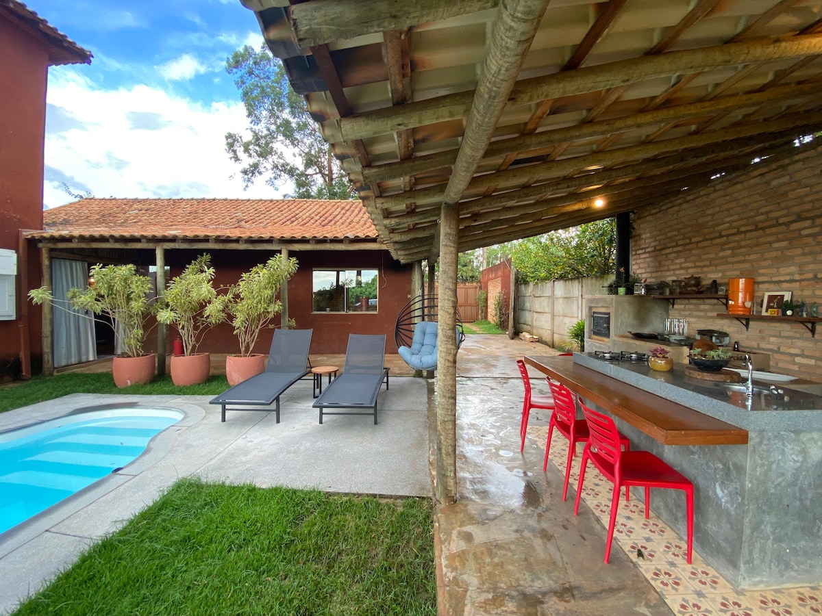 An outdoor area is presented with a swimming pool surrounded by lounge chairs. A covered space features a dining table and chairs, alongside a barbecue station. The setting is complemented by a grassy lawn and a rustic wall made of bricks.