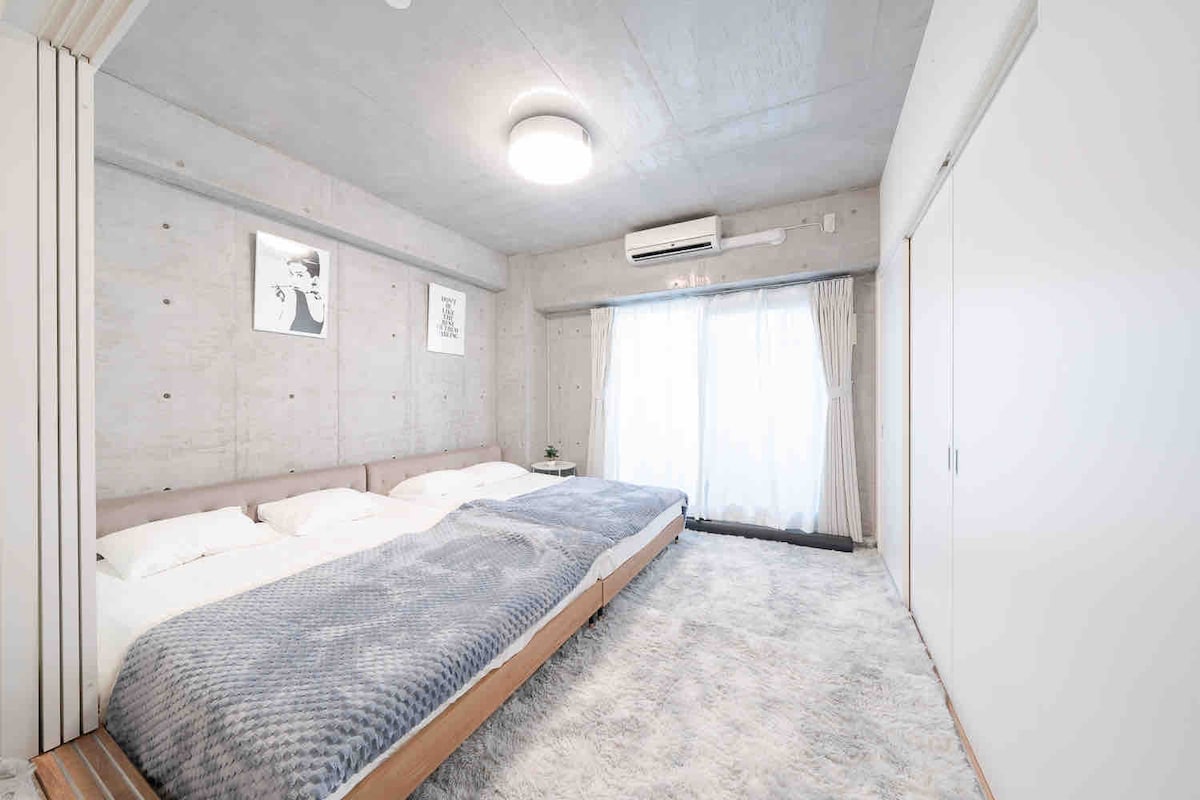 A spacious sleeping area is presented with a light-colored rug and a large bed adorned with soft linens. Two large windows covered by sheer curtains allow natural light to fill the room, highlighting the minimalist decor and concrete walls.