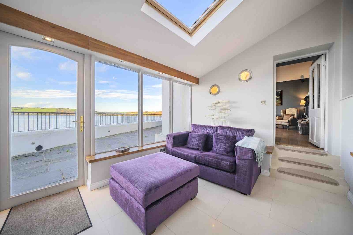 A bright sitting area features large windows allowing natural light to flood the space, showcasing scenic views of the water. A plush purple sofa and matching ottoman provide comfortable seating. Minimalist decor includes wall-mounted lights and soft flooring, enhancing the tranquil ambiance.