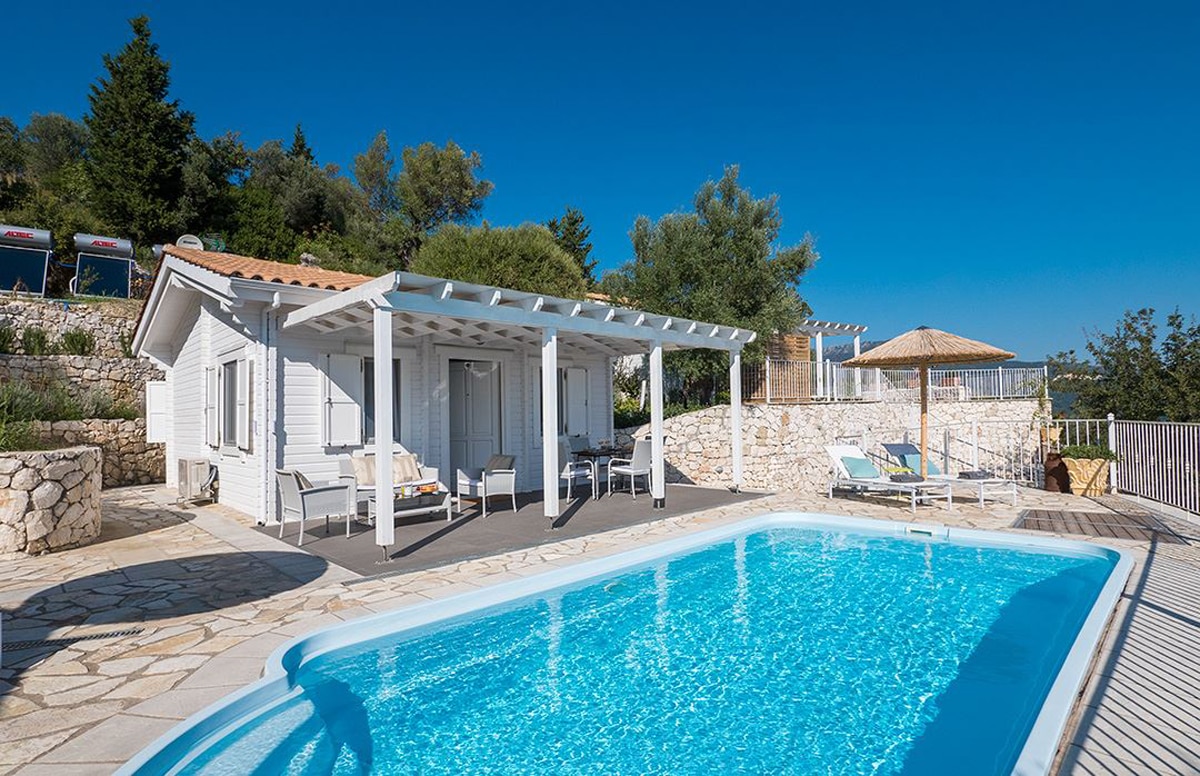 A private pool area features a sparkling blue pool surrounded by a sun terrace. The white exterior of the villa is complemented by a shaded pergola and several lounge chairs, set against a backdrop of green hills and clear blue skies.