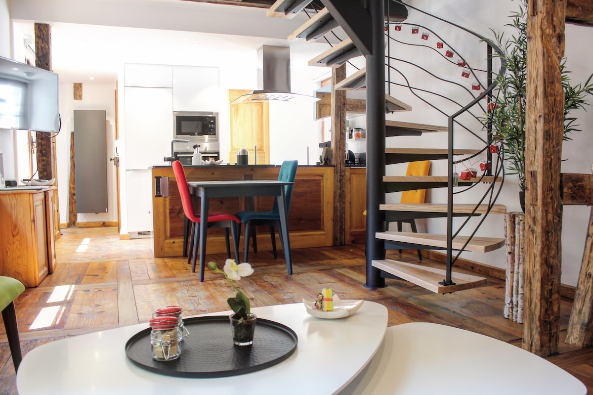 A modern duplex featuring a spiral staircase and an open kitchen with wooden cabinetry. Colorful chairs surround a dining table, while a small decorative tray holds snacks. Natural light illuminates the space, highlighting the blend of rustic wood beams and contemporary design.