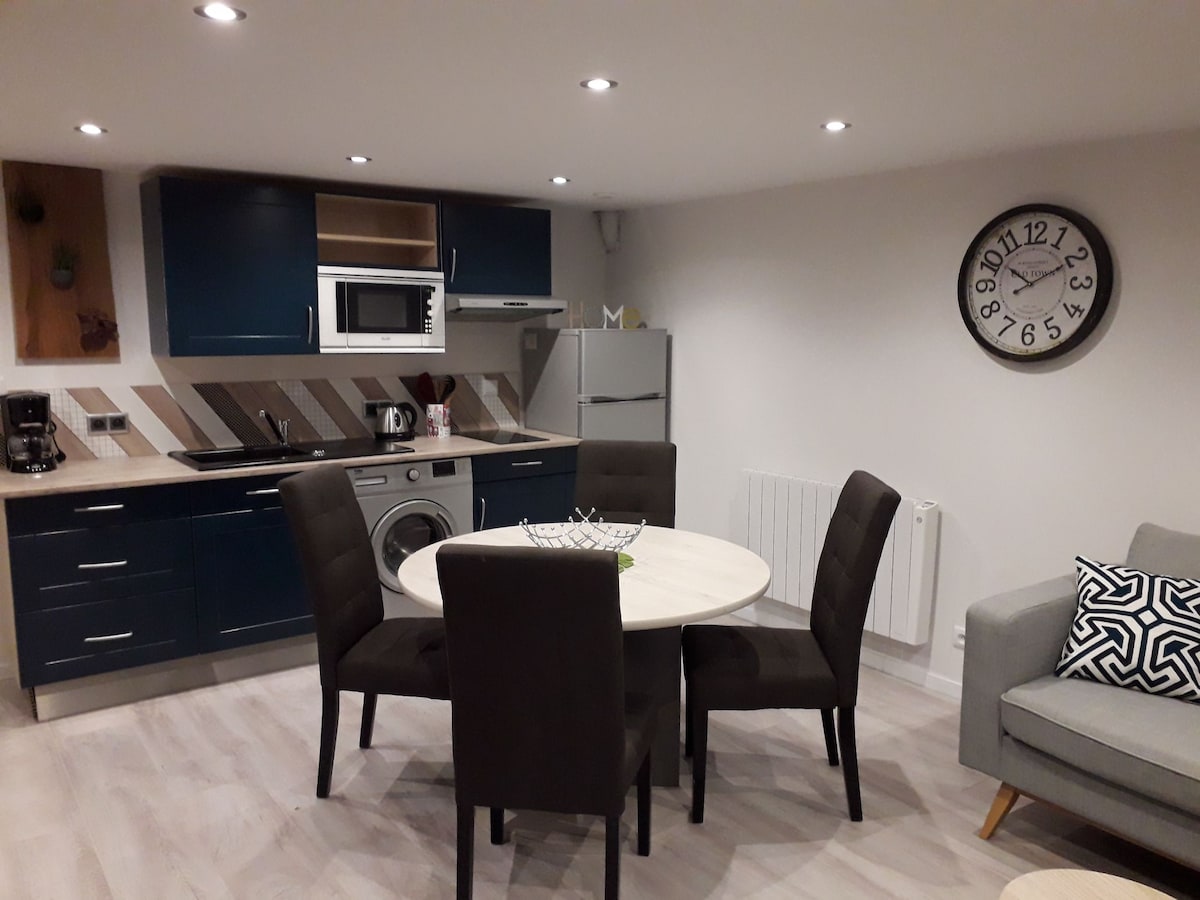 A modern kitchen and dining area feature a circular table surrounded by four dark chairs. An equipped kitchen includes a refrigerator, microwave, and washing machine. Light-colored flooring is complemented by contemporary cabinetry, creating a functional and inviting space. A wall clock adds a practical touch.
