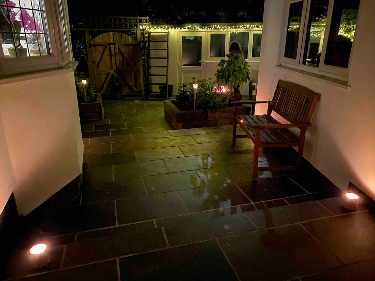 A stone-paved outdoor sitting area is illuminated by soft lighting, featuring a wooden bench positioned along one side. Planters with greenery are arranged around the space, creating a cozy environment. The entrance to a garden area is visible in the background.