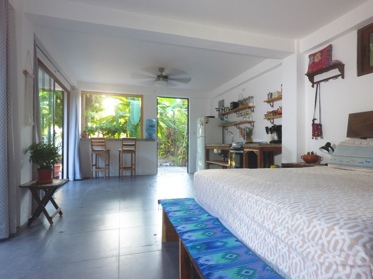 An open living space features a spacious bed and a dining area with barstools. Light filters through large sliding glass doors, illuminating the area. A fully-equipped kitchen is visible alongside a wooden table. A ceiling fan and nearby tropical vegetation enhance the ambiance.