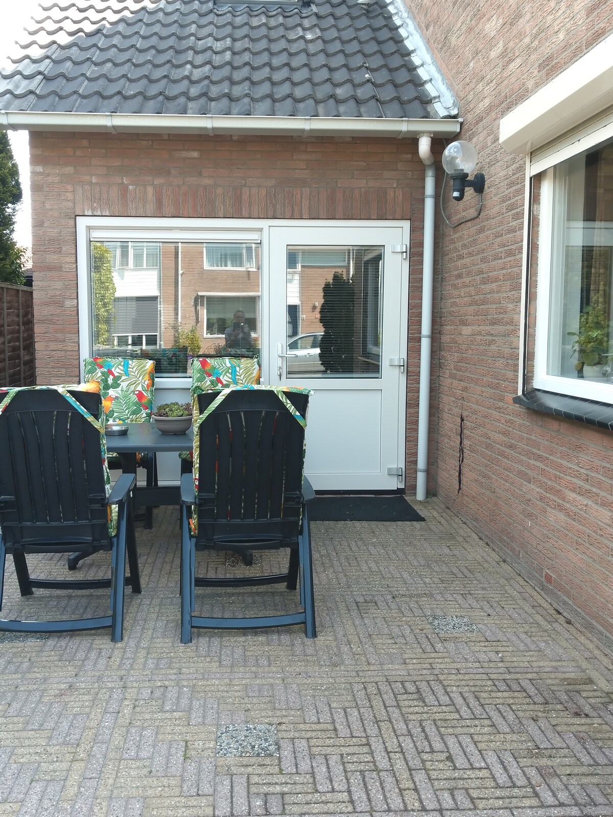An outdoor terrace is displayed, featuring a set of black wooden chairs with vibrant patterned cushions. A small table is positioned in the center, and a door leads into the interior space. The patio is paved with bricks, providing a structured and neat appearance.