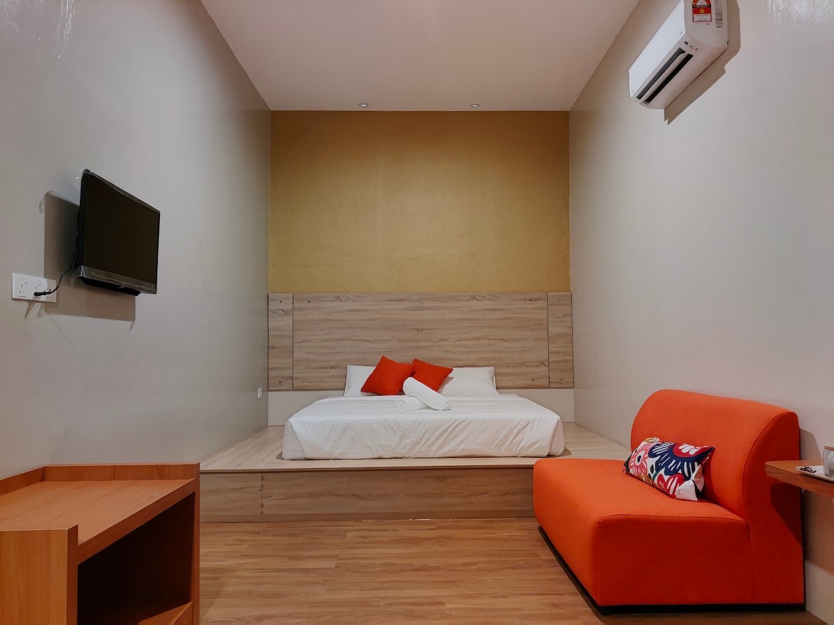 A spacious room is featured with a raised platform bed adorned with white linens and orange accent pillows. An orange sofa sits to the side, and a flat-screen TV is mounted on the wall. Air conditioning is available, with wooden flooring throughout.