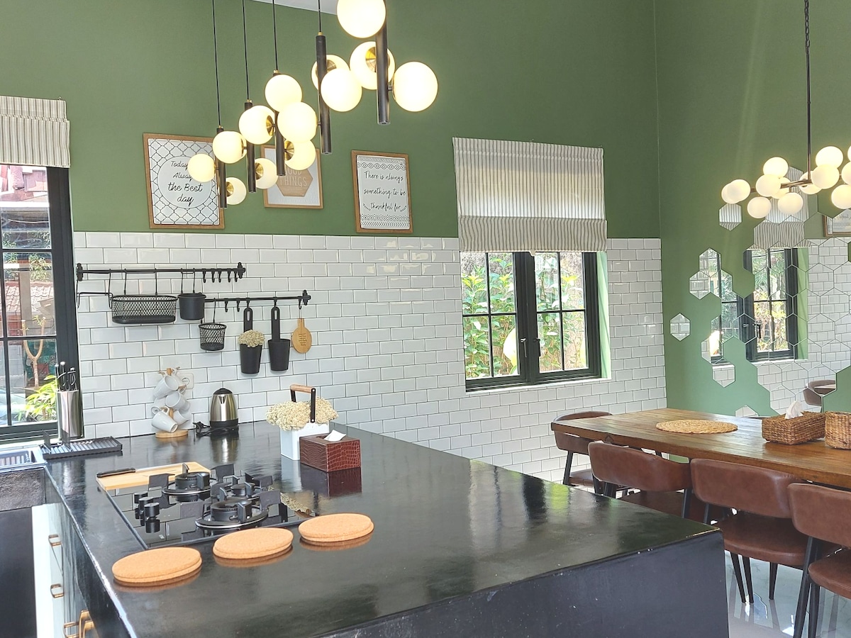 A modern kitchen is highlighted by a sizable island featuring a three-burner gas stove. Light fixtures hang above, and a mix of open shelving and cabinets displays utensils and decor. Large windows allow natural light to illuminate the space, complemented by a rich green wall.