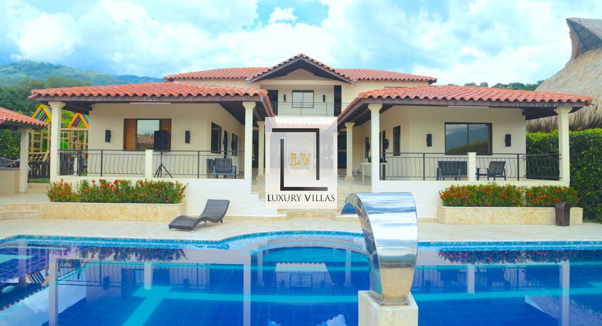 A two-story villa is prominently displayed with a terracotta roof. The pool reflects the structure's exterior, while lounge chairs are positioned nearby. Lush greenery frames the property, enhancing the scenic backdrop of mountains in the distance under a partly cloudy sky.
