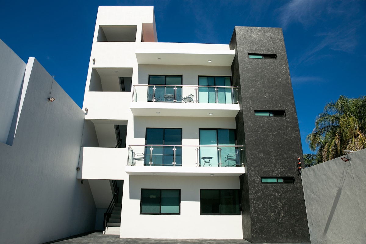 A modern three-story building displays a combination of white and dark exterior walls. Large windows with balconies are positioned on each floor, providing ample natural light. A clear blue sky serves as the backdrop, highlighting the structure's contemporary design.