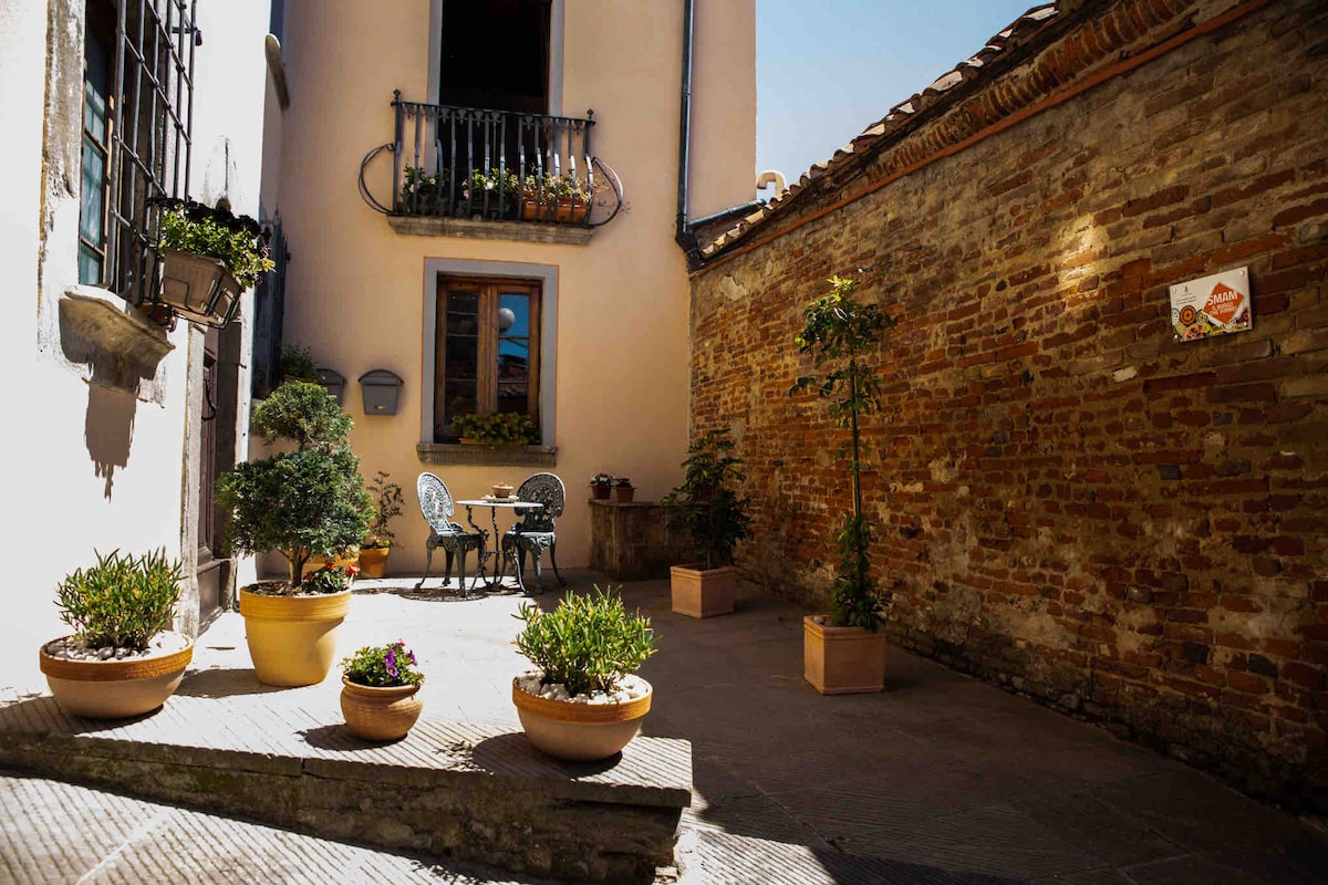 A charming courtyard is featured, defined by a warm brick wall and adorned with potted plants. A small bistro table and two chairs are placed under a balcony, offering a cozy spot for relaxation in the sunshine.