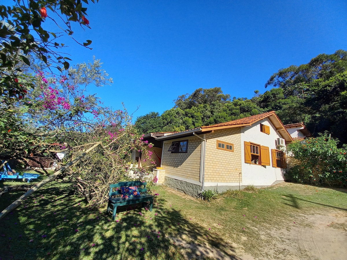 A charming house is nestled among lush greenery, with flowering plants providing vibrant colors. The structure features wooden shutters and air conditioning units on the exterior. A grassy area is visible, offering space for relaxation and outdoor activities.