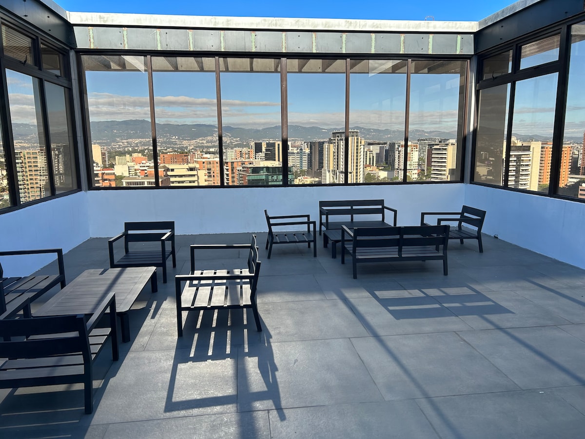 A spacious rooftop lounge area is depicted, featuring multiple black outdoor seating arrangements. Large floor-to-ceiling windows offer panoramic views of the city and surrounding mountains, while ample natural light creates an open atmosphere.