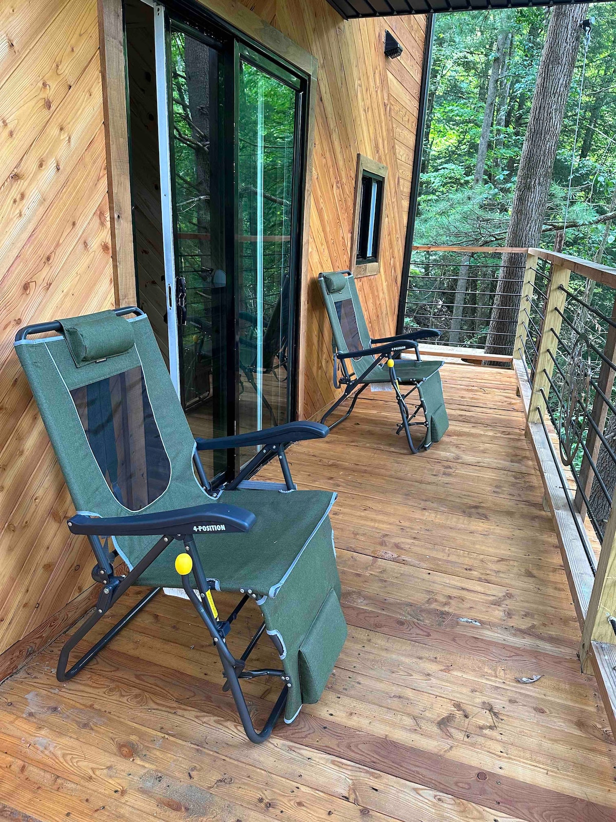 Two comfortable folding chairs are positioned on a wooden deck, offering a view of the surrounding forest. The deck features a railing made of wood and metal, creating a seamless transition between indoor and outdoor spaces. Large glass doors lead into the tree house.