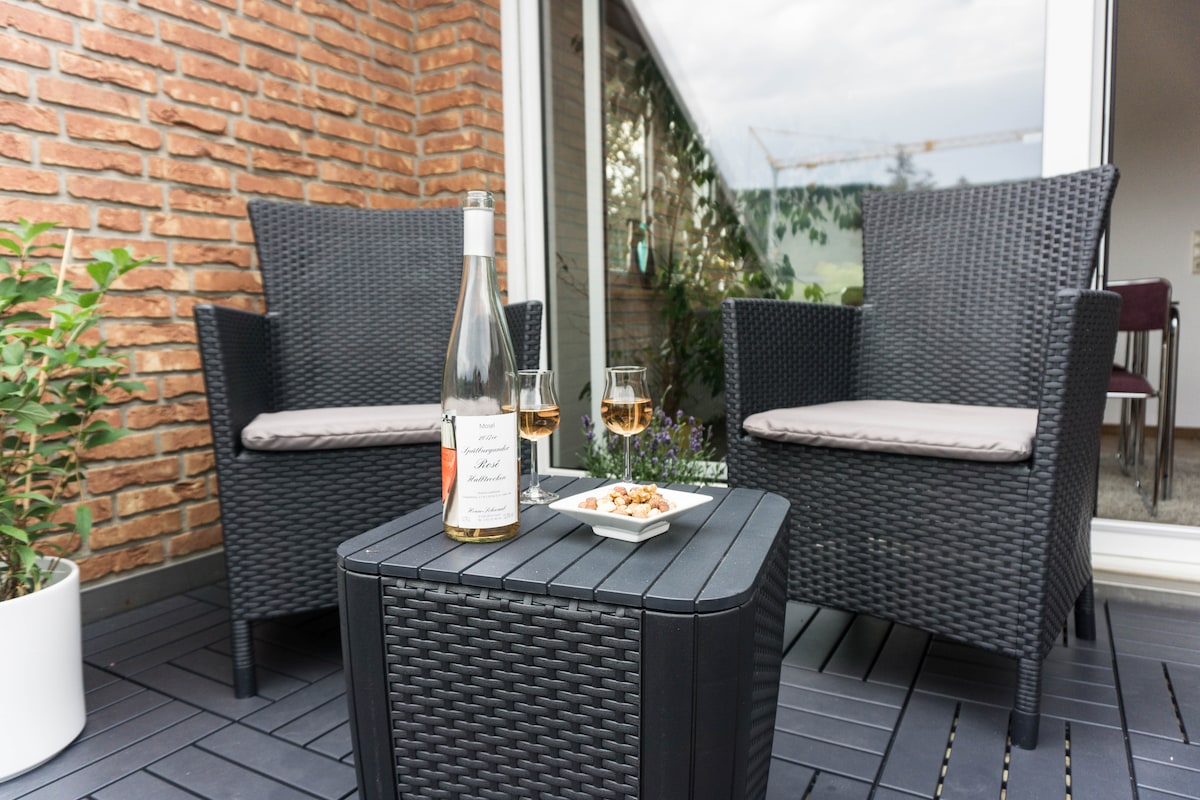 A small outdoor seating area is depicted, featuring two wicker chairs with light cushions surrounding a low table. On the table, a bottle of wine is accompanied by two glasses and a plate of snacks. Verdant plants are visible in the background.
