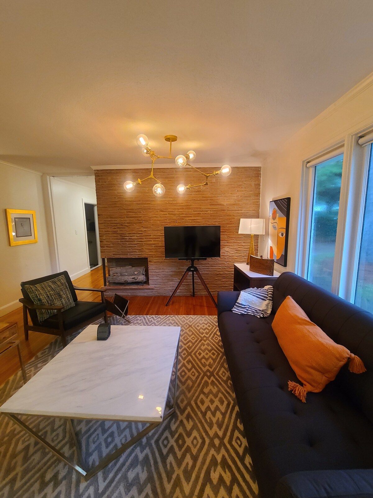 The living room features a sleek sofa bed with accent pillows, complemented by a modern geometric coffee table. A flat-screen TV is mounted against a textured brick wall, and an armchair adds stylish seating. Natural light filters through large windows, enhancing the space.