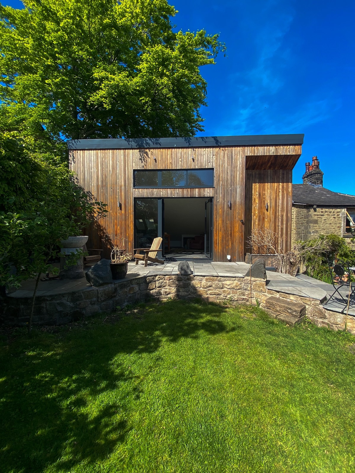 A modern garden room features a wooden exterior, blending naturally with the surrounding greenery. Large windows allow natural light to fill the interior, while a stone path leads to the entrance. Lush grass and potted plants enhance the inviting outdoor space.
