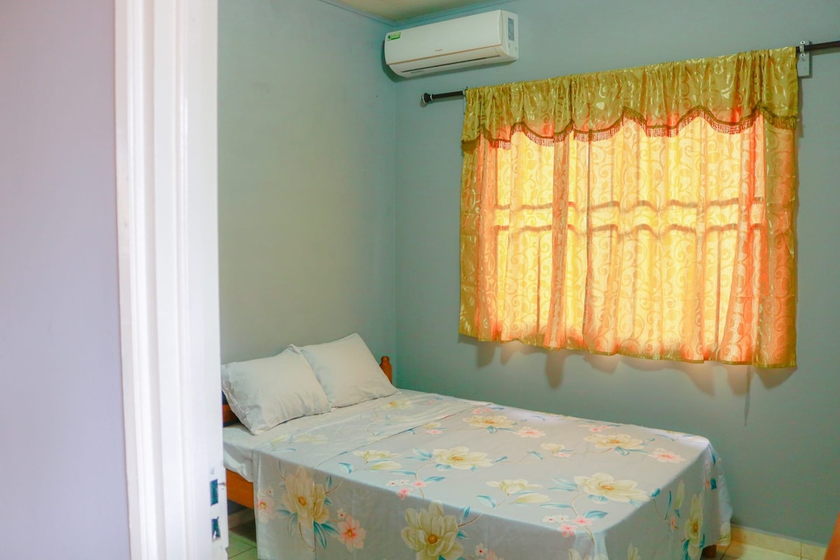 A simple bedroom features a bed covered with a floral sheet and two pillows. Light is filtered through curtains with a warm golden hue, while an air conditioning unit is mounted on the wall, providing a comfortable environment.