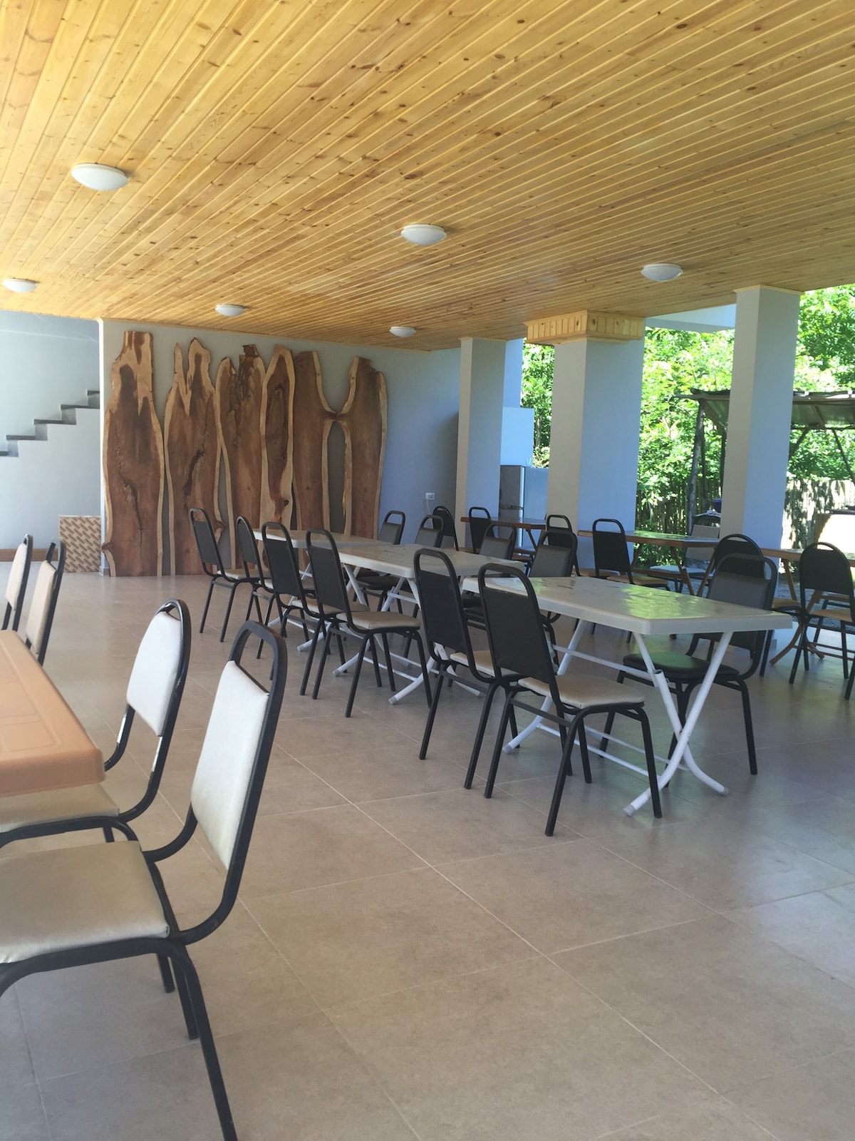 A spacious dining area is presented with numerous metal and plastic chairs arranged around large tables. Natural wood elements are featured prominently on the walls, and a staircase leads to an upper level, enhancing the openness of the space.