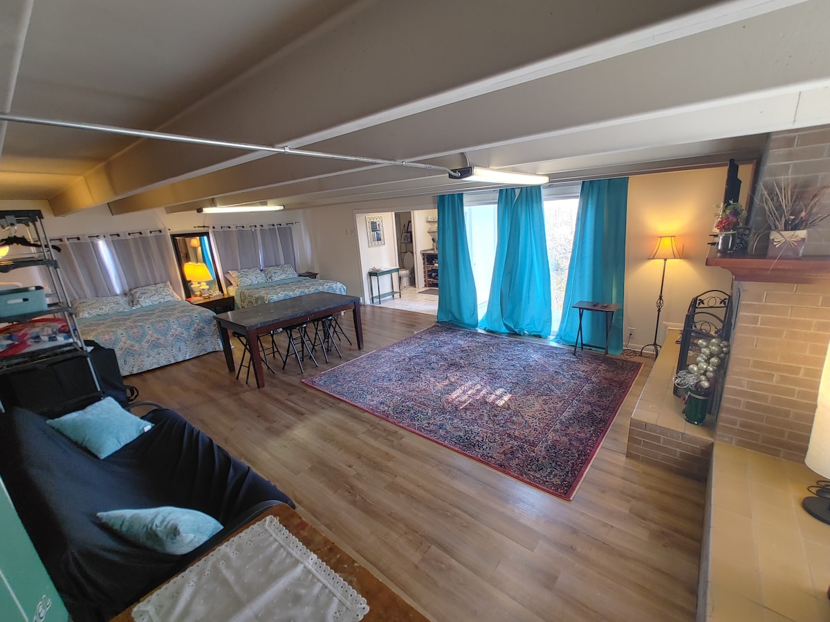An open studio space features two queen beds with patterned bedding and a full futon. A dining table stands center, surrounded by a large area rug. Natural light enters through floor-length turquoise curtains, creating a warm ambiance against the neutral walls.