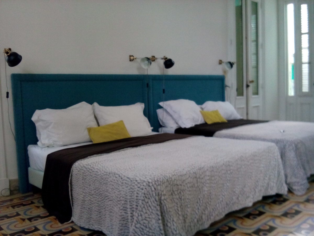 A spacious room features two large beds adorned with soft linens. One bed is dressed in a light coverlet, while the other is topped with a dark brown throw. Bedside lamps provide focused lighting, complementing the bright atmosphere enhanced by natural light from the windows.