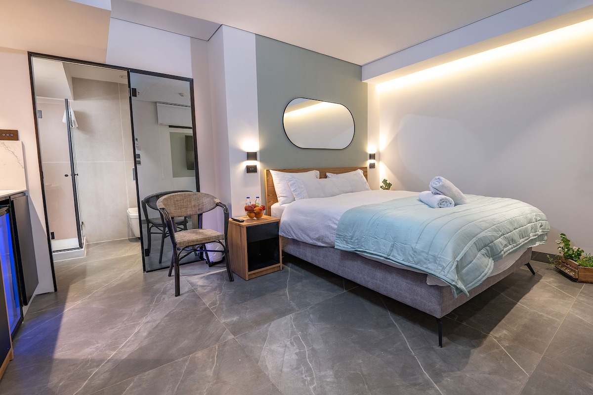 A well-appointed bedroom features a spacious bed with a light blue blanket and fresh towels. A round mirror is mounted above a bedside table. A large window allows natural light to fill the room, which is complemented by a stylish chair and small fruit bowl.