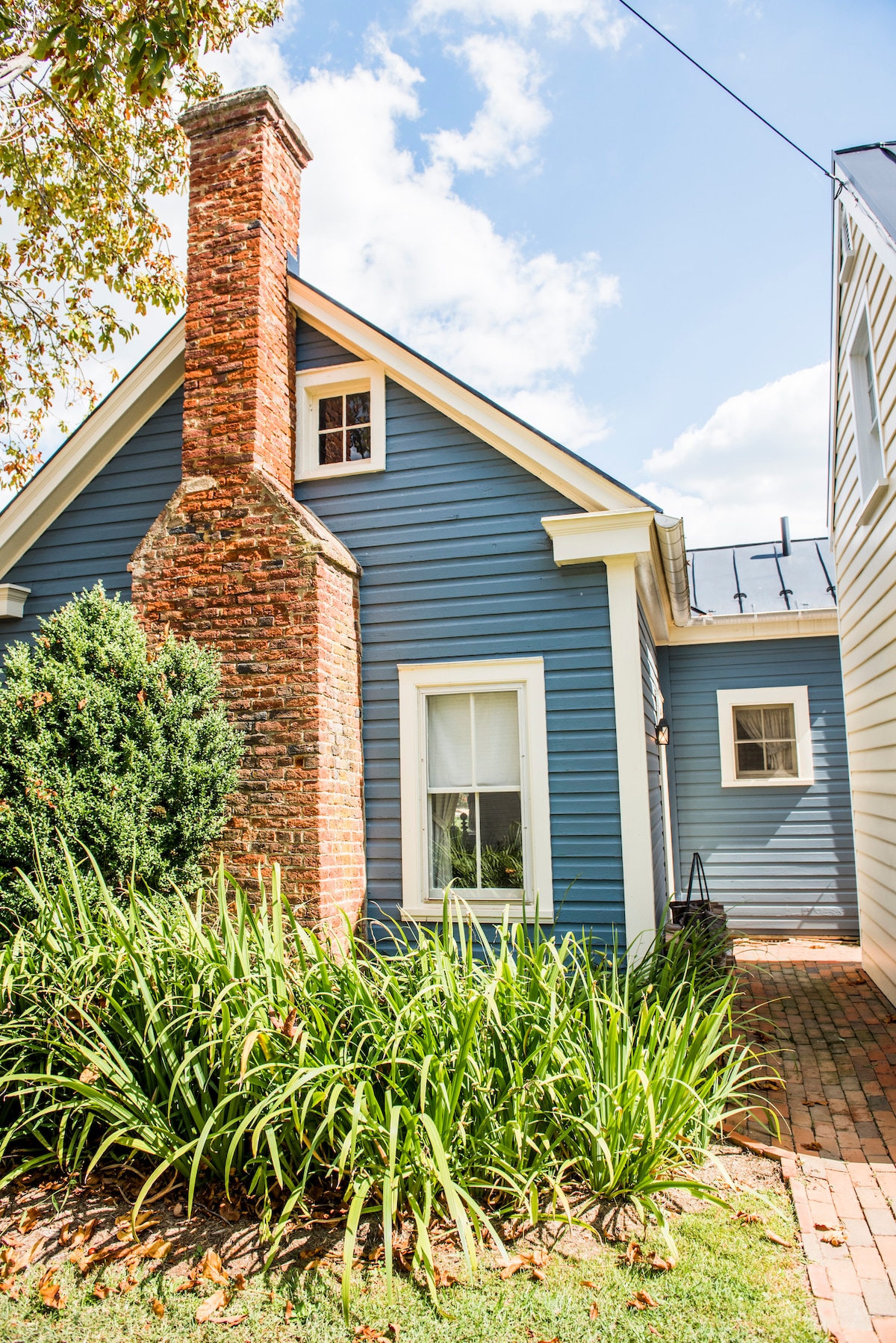 The exterior of the Patent House is showcased, featuring a blue facade complemented by a brick chimney. Lush green plants surround the base of the home, with a few steps leading up to the door and a clear sky above.