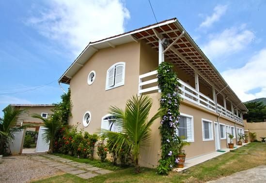A multi-unit building features a two-story design with white accents and a terrace lined with plants. The exterior walls are painted in a light beige tone. Paths with stones provide access, and lush greenery is visible throughout the property.