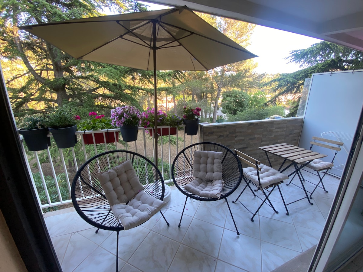 A balcony area features two cushioned chairs and a small foldable table, shaded by a large umbrella. Green plants and flower pots line the railing, enhancing the serene outdoor setting overlooking a garden.