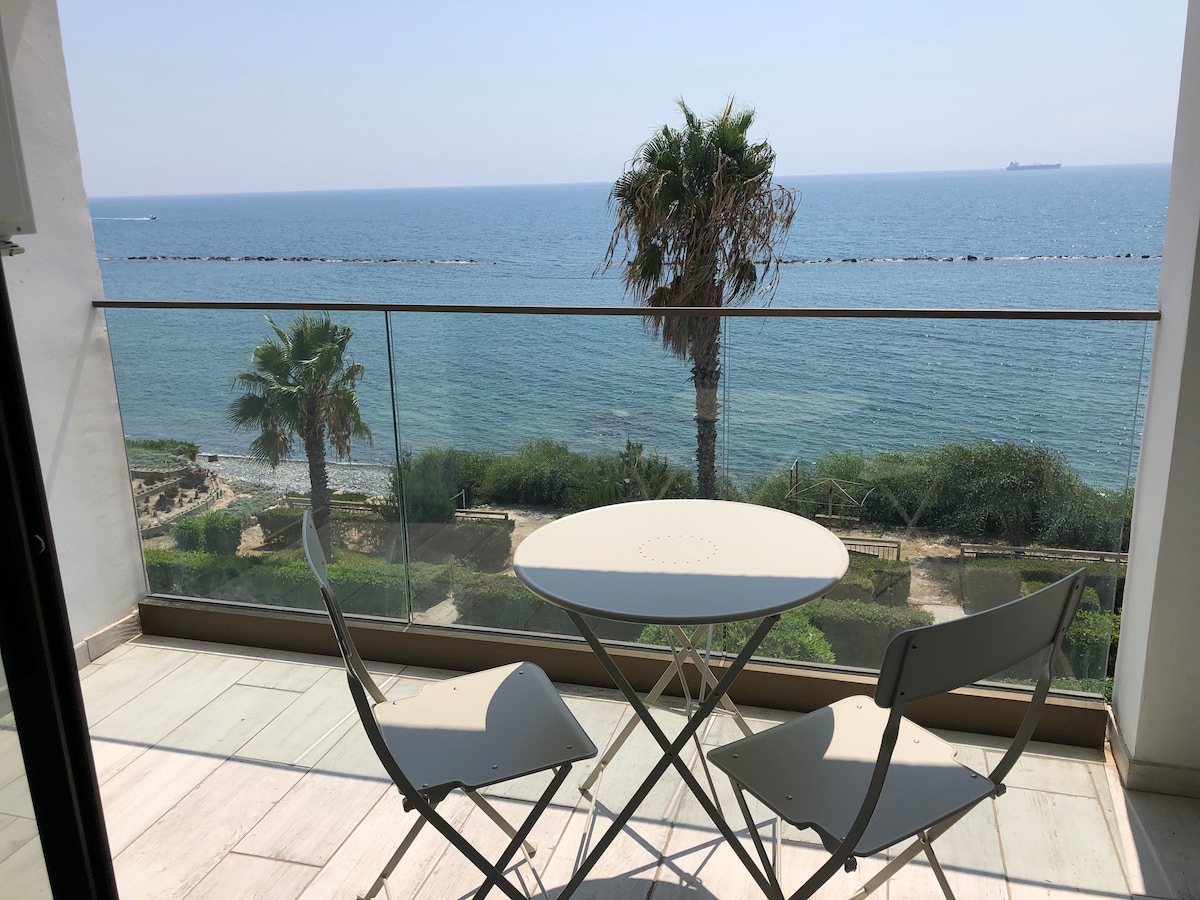 A balcony offers a direct view of the open sea, with shimmering blue waters stretching to the horizon. A small round table and two chairs provide a space for relaxation. Lush greenery and palm trees frame the seafront in the background.