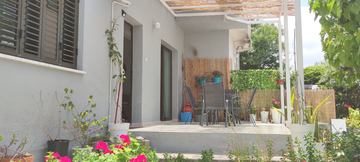 A welcoming outdoor patio features a sturdy table surrounded by chairs, set under a covered area. Potted plants and vibrant flowers add greenery to the space, enhancing the serene environment. The entrance can be seen leading into the living area.