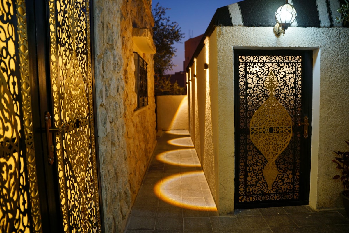 A softly lit pathway is framed by decorative metalwork gates, leading to private entrances. Warm lighting accentuates the stone wall and tiled flooring, creating a welcoming ambiance. The path is bordered by greenery, enhancing the tranquil setting as twilight approaches.
