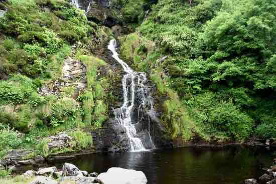 A serene waterfall cascades down rocky terrain, surrounded by lush greenery. The water flows into a dark pool, reflecting the surrounding foliage and rocks. The vibrant vegetation contributes to a tranquil natural setting, inviting peaceful contemplation and exploration.