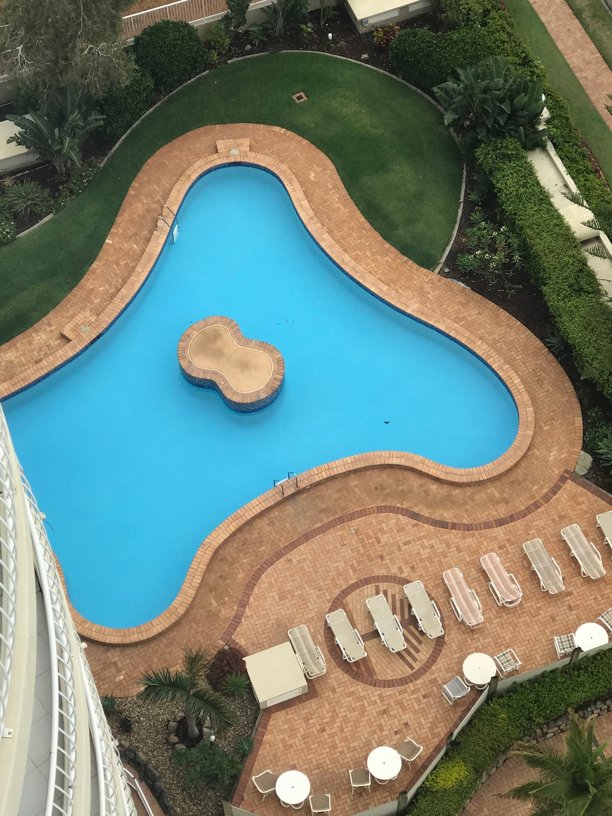 An aerial view of the outdoor pool area is presented, featuring a uniquely shaped pool surrounded by a tiled deck. Several lounge chairs and tables are positioned nearby, complemented by well-kept greenery and landscaping that enhances the relaxing atmosphere.