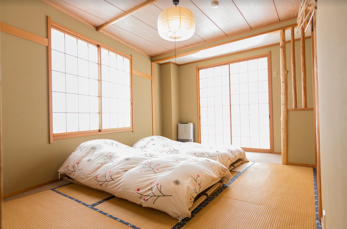 A Japanese-style room is presented with tatami flooring and two futons laid out. Natural light filters through large shoji windows, creating a calm and serene environment. A simple light fixture hangs from the ceiling, and a small heater is visible in the corner.